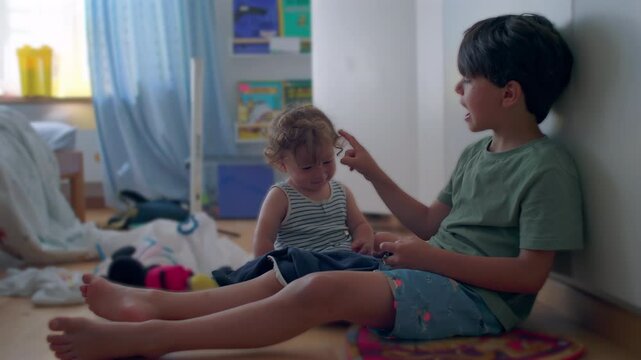 Older brother playfully teases toddler by tapping face repeatedly while sitting together on bedroom floor, lighthearted sibling moment inside child’s room