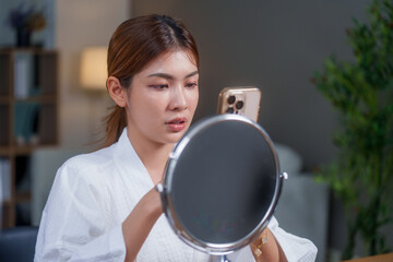 Woman in bathrobe applies makeup while looking at her phone and mirror, showcasing her beauty...