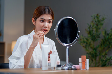 Woman in bathrobe applies makeup in front of mirror, showcasing her beauty routine with various...
