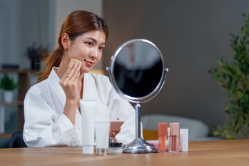 Woman in bathrobe applies makeup while looking in mirror, surrounded by various cosmetics. Her...