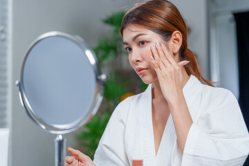 Woman in bathrobe applies makeup while looking in mirror, showcasing her beauty routine with...