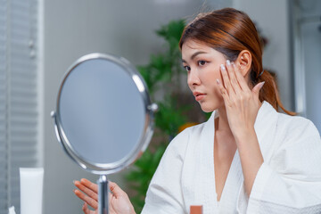 Woman in bathrobe applies makeup and cosmetics while looking at her reflection in mirror,...