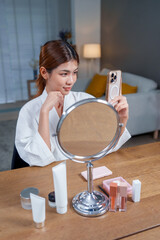 Woman in bathrobe is applying makeup while looking at her phone in front of mirror. scene captures beauty and cosmetics, showcasing various products on wooden table
