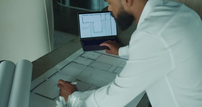 An architect is currently analyzing and working on building plans using a laptop and blueprints