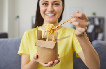 Happy woman eating pasta from takeout carton box at home. Young person holding paper container and...