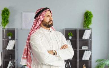 Thoughtful Arabian man wearing ghutra on head, keeping arms folded, looking aside pensively, being deep in thoughts, contemplating about something, posing against office interior. 