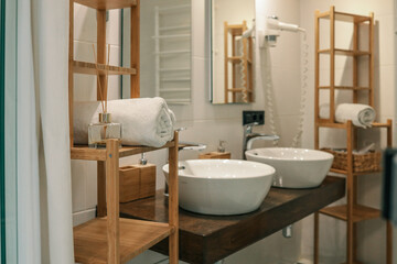 Rolled white towel and reed diffuser on a wooden shelf in a modern bathroom with double sinks and...