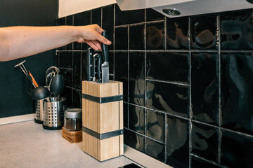 Hand reaching for a kitchen knife from a wooden knife block on a modern countertop with black tile...