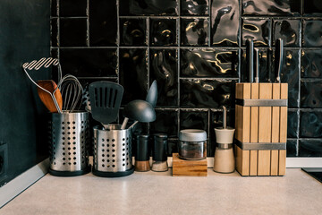 Modern kitchen countertop with utensils, spice jars, a wooden knife block, and black glossy tile...