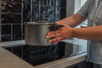 Person placing a metal pot on an induction stove in a modern kitchen with black tile backsplash and...