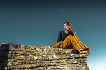 Young woman sitting in a sleeping bag on a rocky mountain cliff at sunset, gazing into the distance with the moon in the sky