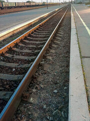 Curved railway tracks at a quiet train station during sunset. The scene features multiple railway lines, overhead electric wires, and a deserted platform with a few distant pedestrians. The warm