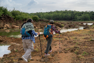 Two people are walking on a dirt path near a river. One of them is wearing a plaid shirt