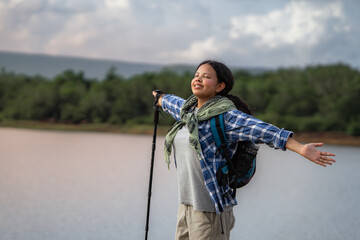 A woman is standing on a bridge with a backpack and a walking stick. She is smiling and looking at the water