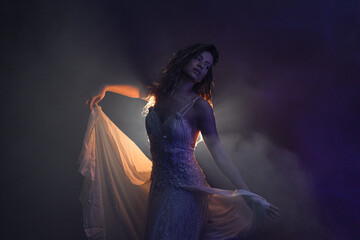 A beautiful young woman in a white dress dances in a haze with a mysterious light from behind,...
