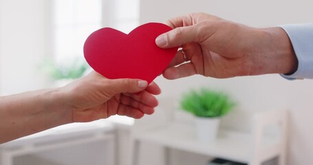 Close up of two people hands exchanging and holding red paper heart, loving man giving heart to woman. Symbolic gesture of love, romance, care, kindness, charity, support and emotional connection.
