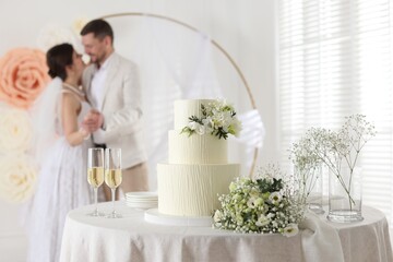 Happy married couple together indoors, focus on wedding cake, glasses of champagne and bouquet on table