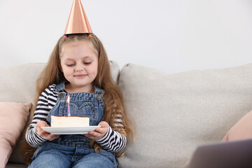 Birthday celebration. Little girl with piece of cake having video call via laptop on sofa at home
