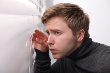 Paranoia. Worried man looking through window blinds indoors