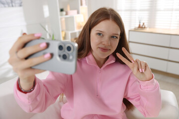 Beautiful teenage girl taking selfie by smartphone and showing peace sign on sofa at home