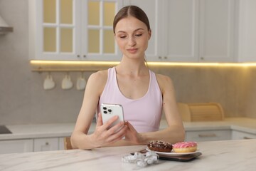 Woman using smartphone at white marble table with kitchen scale, donuts and measuring tape indoors