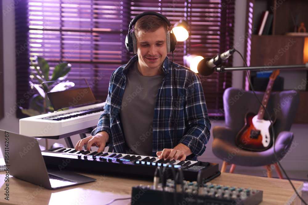 Poster smiling man in headphones working with midi keyboard at table in home studio