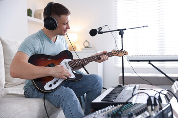 Man in headphones working with guitar and MIDI keyboard on sofa in home studio