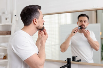 Handsome man shaving with trimmer near mirror at home, selective focus