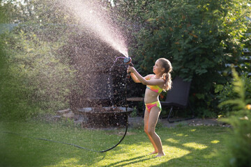Happy little  girl playing with garden hose and having fun with spray of water in sunny backyard....