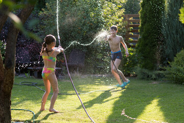 Happy children brother and sister playing in the garden hose and having fun with spray of water in...