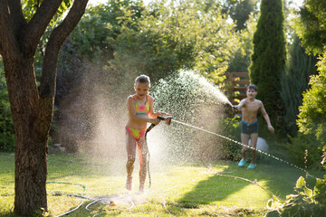 Happy children brother and sister playing in the garden hose and having fun with spray of water in...