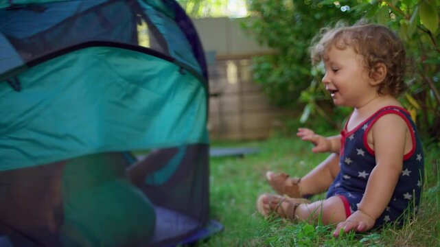Laughing baby crawls toward play tent in sunny backyard as older brother hides inside and rattles fabric, creating a joyful and carefree moment of sibling fun - Powered by Adobe