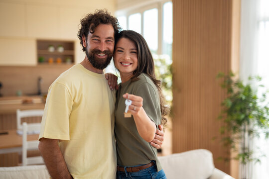 Happy couple smiling and holding a house key in their new home, standing close together in a bright living room.
