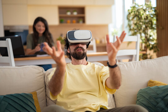 Man using VR headset on the sofa, interacting with virtual content. Woman in the background working on a laptop at the kitchen table.