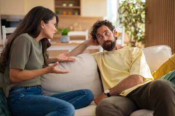 Obraz premium Young couple having a disagreement while sitting on a sofa at home. The woman gestures emotionally while the man listens with a tired or dismissive expression, suggesting tension in the relationship. 