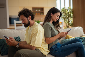 Young couple sitting back to back on a sofa, both absorbed in their smartphones. The image reflects...