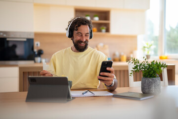 Smiling man wearing headphones, using a smartphone while sitting at a kitchen table with a tablet and notebook.