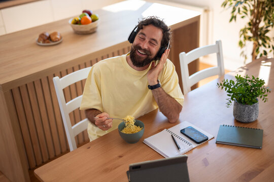 Happy man wearing headphones and enjoying a meal while sitting at the table with a notebook and tablet at home.
 - Powered by Adobe