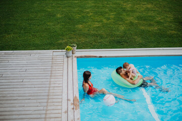 Young family with small daughter in swimming pool outdoors in backyard garden.
