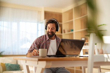 Smiling man wearing headphones and reaching for a coffee mug while working on a laptop in a bright home office.
