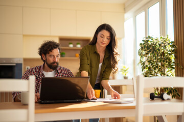Young couple working together at home, man using a laptop while woman organizes papers on the kitchen table.
