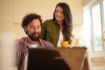 Young couple smiling and spending time together at home, man working on a laptop while woman stands beside him holding a mug.
