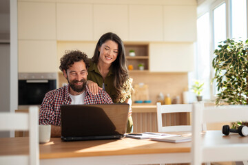 Young couple smiling while looking at a laptop together in a cozy kitchen, enjoying a relaxed moment at home.
