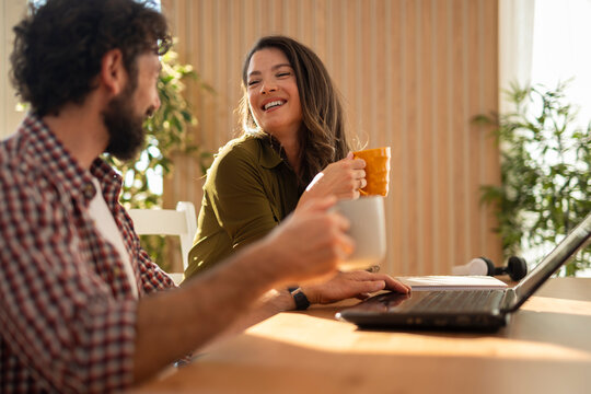 Happy couple enjoying coffee together at home, smiling and chatting at the table with a laptop in front of them.
