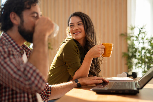 Happy young couple sitting together at home, woman smiling while holding a mug and man drinking coffee beside a laptop.
