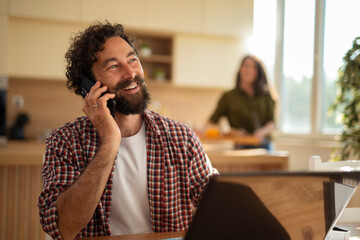 Cheerful man talking on the phone while working on a laptop at home, with a woman in the background...