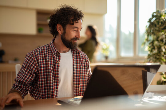 Bearded man working on a laptop at home, looking to the side with a woman blurred in the background in a bright kitchen.
 - Powered by Adobe