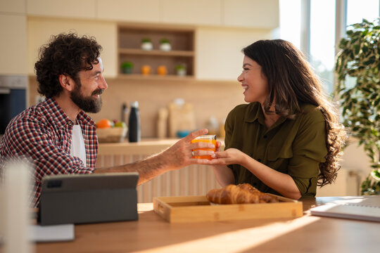 Happy couple having breakfast together at home, woman handing a glass of orange juice to man, enjoying a cozy morning with croissants on a tray and digital tablet nearby.
- Powered by Adobe
