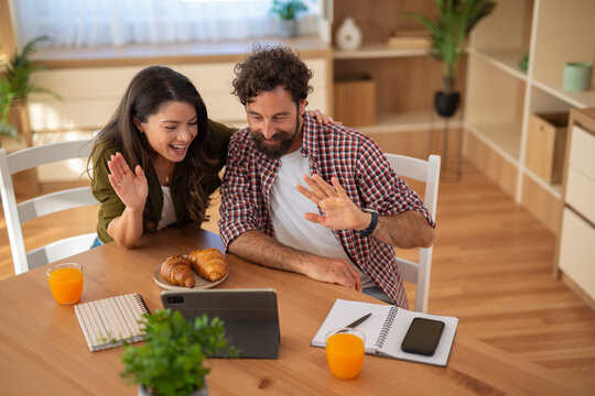 Young couple video calling on tablet while sitting at home kitchen table.