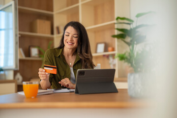Fototapeta premium Woman with credit card using tablet at kitchen table, smiling and relaxed. 